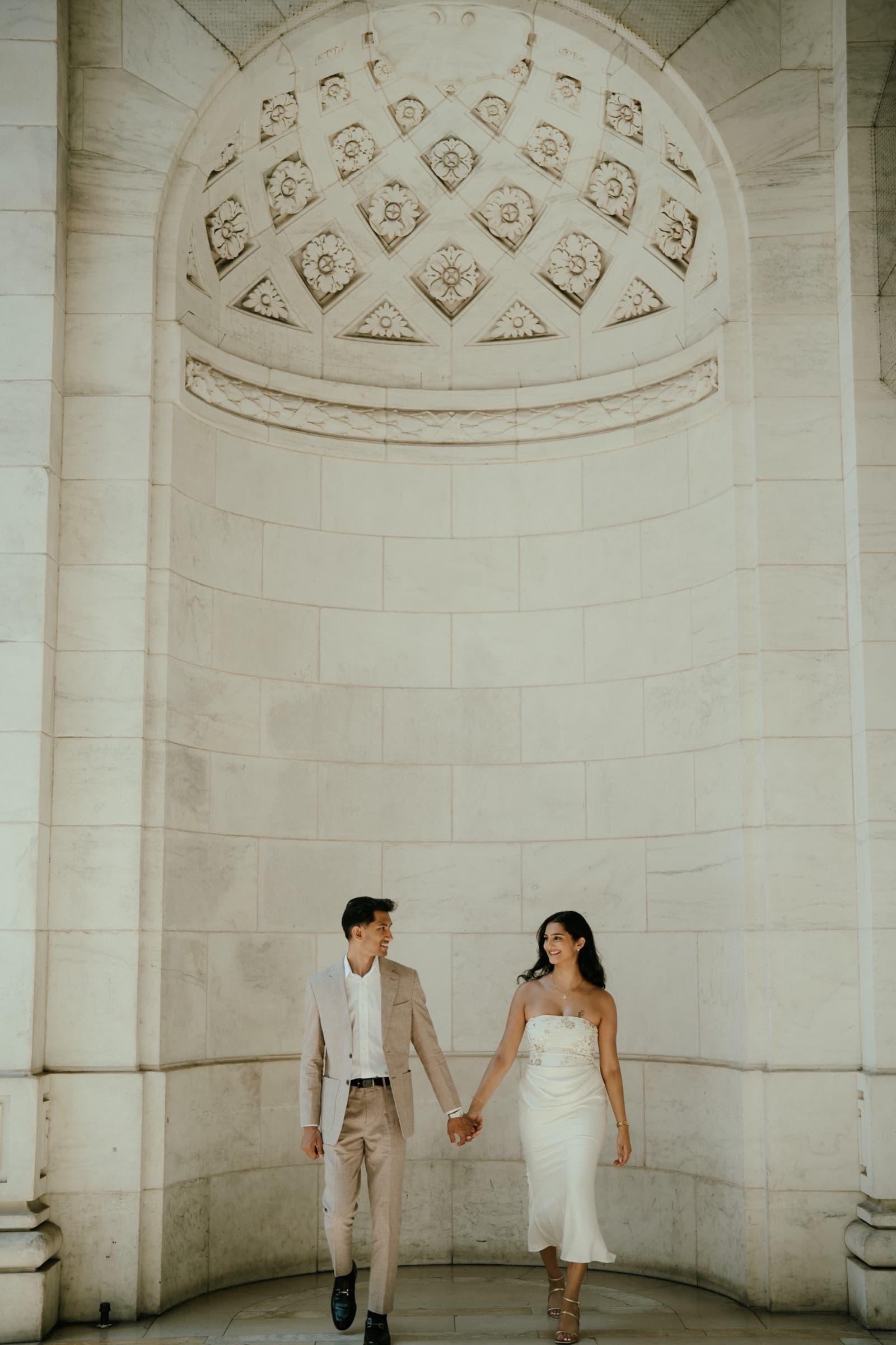 Ananya and Varun walking hand in hand beneath a carved stone arch
