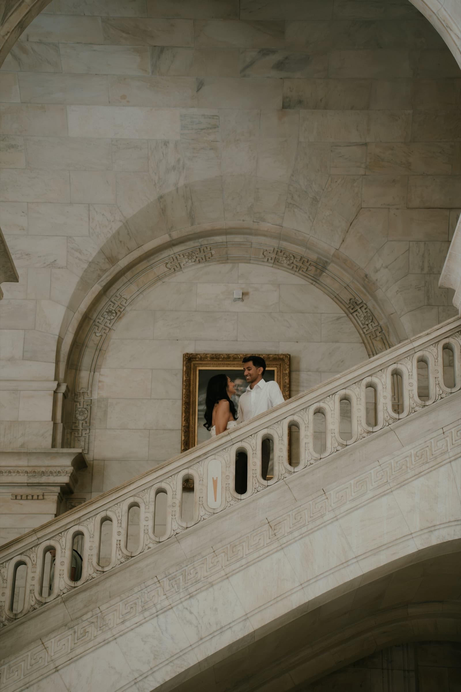 Ananya and Varun smile at each other on a landing framed by a grand marble arch and staircase